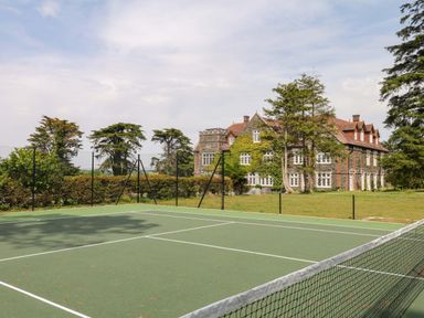 A tennis court in front of a large house surrounded by trees at Alston Hall near Mothecombe Beach