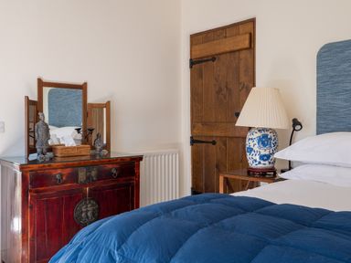 A bedroom with a blue bedcover wooden door a bedside table with a lamp and a wooden dresser with a mirror and decorative figurines at Bank Barn Wreay Watermillock