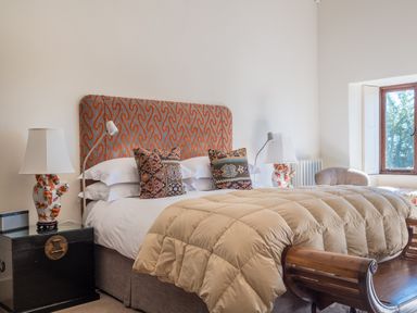 A bedroom with a bed with patterned headboard two bedside tables with lamps a window and a chair at Bank Barn Wreay Watermillock