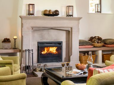 A living room with a lit fireplace armchairs a glass table with glasses and a bowl of fruit at Bank Barn Wreay in Watermillock
