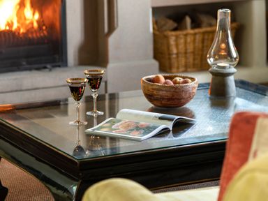 A glass coffee table with two glasses of red wine an open book a bowl of fruit and a candle in a living room with a fireplace at Bank Barn Wreay in Watermillock