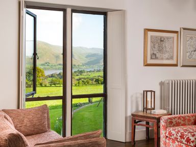 A seating area with patterned armchairs and a wooden side table beside a large open window with a view of green fields and hills at Bank Barn Wreay in Watermillock