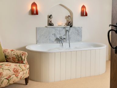 A bathtub with running water and candles on a shelf above in a bathroom with a floral armchair nearby at Bank Barn Wreay in Watermillock