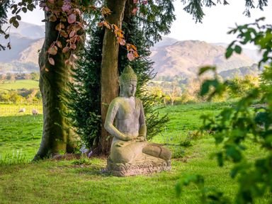 A stone Buddha statue sitting on grass near trees with mountains and sheep in the background at Bank Barn Wreay Watermillock