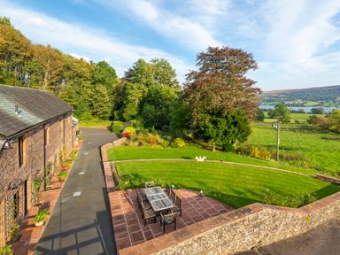 An outdoor patio with a table and chairs next to a stone building and a curved lawn with trees and fields in the background at Bank Barn Wreay in Watermillock