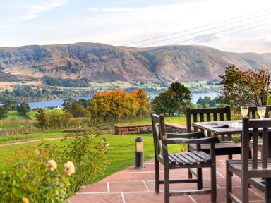 An outdoor patio with wooden chairs and a table overlooking a grassy field with trees a lake and mountains at Bank Barn Wreay in Watermillock