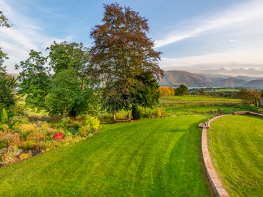 A large green lawn with trees and a curved stone wall overlooking fields and hills at Bank Barn Wreay Watermillock