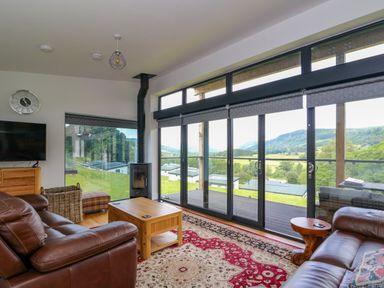 A living room with brown leather sofas a coffee table a wood stove and large windows overlooking a green landscape at 6 Strathtay Lodges in Aberfeldy