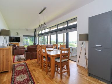 A dining table with six wooden chairs and a living area with leather sofas and a television in a room with large windows at 6 Strathtay Lodges in Aberfeldy