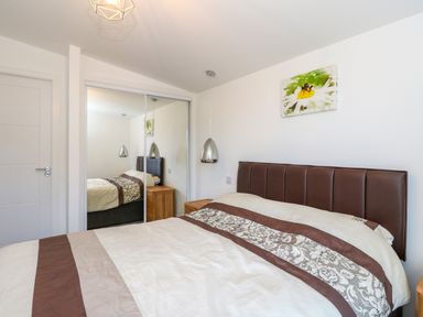 A bedroom with a bed with brown and beige bedding a bedside table with a pendant lamp and mirrored closet doors at 6 Strathtay Lodges in Aberfeldy