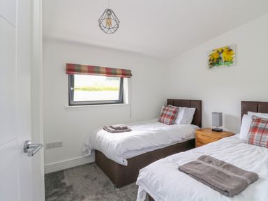 A bedroom with two single beds with white bedding and plaid pillows a wooden nightstand with a lamp and a window with a striped blind at 6 Strathtay Lodges Aberfeldy