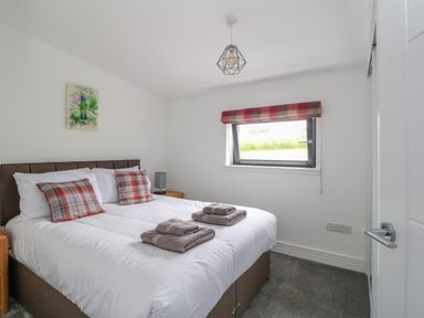 A bedroom with a bed with plaid pillows and folded towels on it a window with a plaid valance a bedside table a small lamp and a painting on the wall at 6 Strathtay Lodges in Aberfeldy