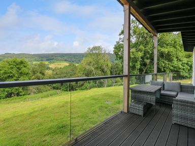 An outdoor deck with a wicker chair and table set overlooking a grassy hillside and tree-covered hills at 6 Strathtay Lodges Aberfeldy