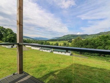 A view of green fields and hills from a balcony with wooden post and glass railing at 6 Strathtay Lodges in Aberfeldy