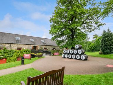 A stone building with a slate roof surrounded by green grass and trees with stacked barrels spelling home of dewars in Aberfeldy