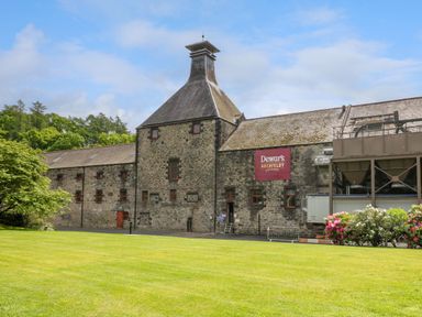 An old stone distillery building with Dewars Aberfeldy sign and green lawn in front at 6 Strathtay Lodges in Aberfeldy