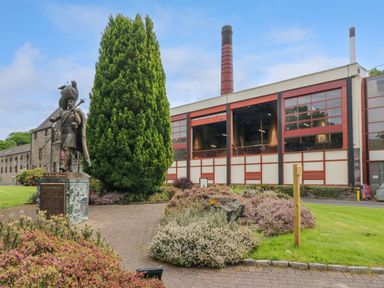 A statue of a bagpiper near tall tree and industrial building with a chimney at 6 Strathtay Lodges in Aberfeldy