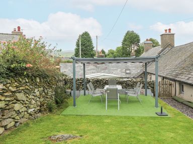 An outdoor seating area with a table and six chairs under a metal pergola in a garden with stone walls at Penrallt in Rhydlydan