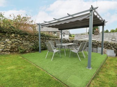 An outdoor seating area with a table and six chairs under a pergola on artificial grass at Penrallt in Rhydlydan