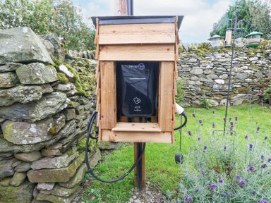 An electric vehicle charging station housed in a wooden enclosure with stone walls and grass in the background at Penrallt in Rhydlydan
