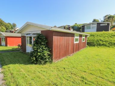 A red wooden cabin with white trim and windows surrounded by grass and nearby similar cabins at Milkwood in Elmrise Park near Llansteffan