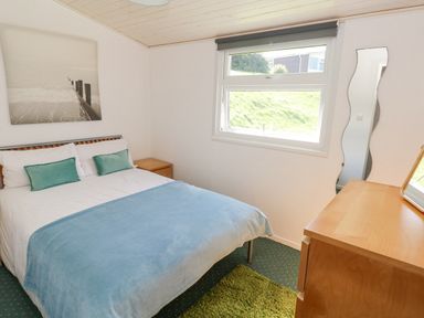 A bedroom with a double bed and blue blanket a wooden dresser and a grassy view at Milkwood in Elmrise Park near Llansteffan