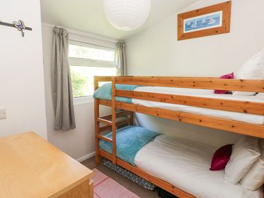 A small bedroom with wooden bunk beds blue blankets white pillows pink cushions a wooden desk a window with grey curtains and a framed nautical picture on the wall at Milkwood in Elmrise Park near Llansteffan