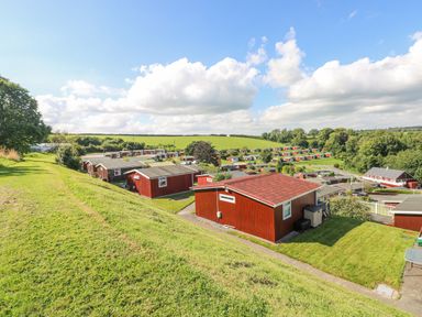 A hillside view of red wooden cabins with green lawns and trees at Milkwood in Elmrise Park near Llansteffan