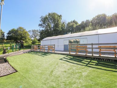 An outdoor area with artificial grass wooden benches and a large white tent with trees in the background at Milkwood in Elmrise Park near Llansteffan