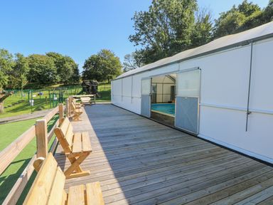 A wooden deck with benches alongside a covered swimming pool and a fenced playground in the background at Milkwood in Elmrise Park near Llansteffan