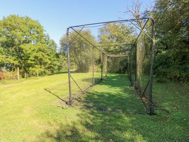 An outdoor cricket practice net with a metal frame and netting on a grassy area surrounded by trees at Milkwood in Elmrise Park near Llansteffan