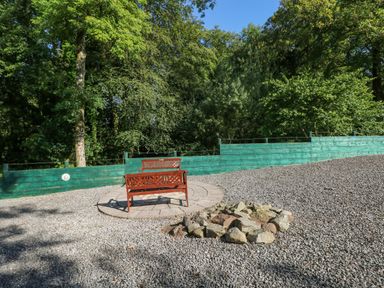 An outdoor area with two wooden benches on a circular paved platform and a fire pit made of stones near a green wooden fence with trees behind at Milkwood in Elmrise Park near Llansteffan