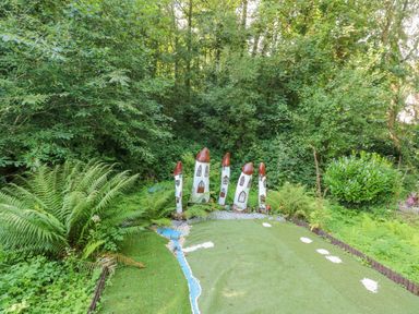 A garden area with green artificial grass and tall mushroom-shaped sculptures surrounded by trees and plants at Milkwood in Elmrise Park near Llansteffan
