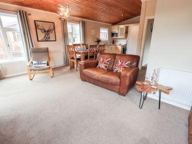 A living room with a leather sofa with British flag pillows a wooden armchair with a cushion a dining table with chairs kitchen appliances and a wooden ceiling at Brightwater Lodge White Cross Bay near Windermere