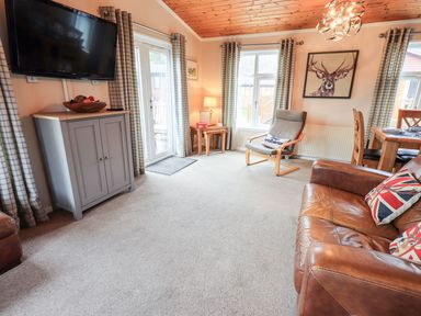 A living room with a brown leather sofa with cushions a wooden armchair a wall mounted television a small cabinet and a dining table at Brightwater Lodge White Cross Bay near Windermere