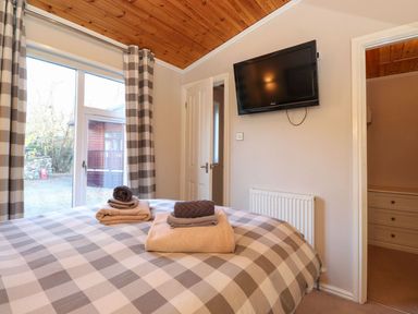 A bedroom with a bed covered in checkered bedding towels on the bed a wall-mounted television a window with checkered curtains and a door leading to another room at Brightwater Lodge White Cross Bay near Windermere