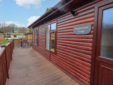 An outdoor deck with wooden railing and a table with chairs outside a wooden lodge named Brightwater Lodge at White Cross Bay near Windermere
