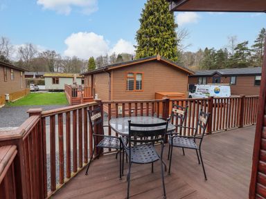 A wooden deck with a glass table and four metal chairs overlooking cabins and trees at Brightwater Lodge White Cross Bay near Windermere