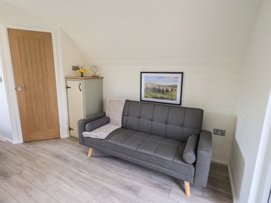 A gray sofa with a blanket and cushions next to a wooden door and a small cabinet with flowers and a heart decoration at Shepherds View in Castlerigg near Keswick