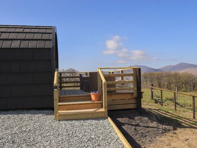 A wooden deck with stairs and a flower pot next to a dark curved structure in a rural area at Shepherds View in Castlerigg near Keswick