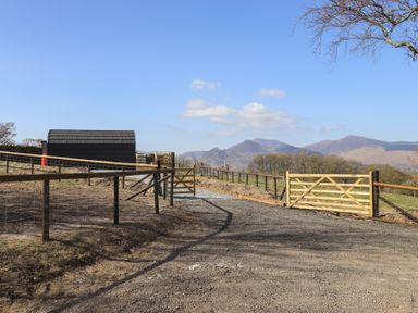 A fenced gravel driveway with wooden gates and a small black shed with mountains in the background at Shepherds View Castlerigg near Keswick