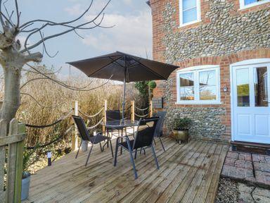 An outdoor deck with a table, four chairs, an umbrella, and a tree next to a stone and brick building at Cobble Cottage in Great Ryburgh
