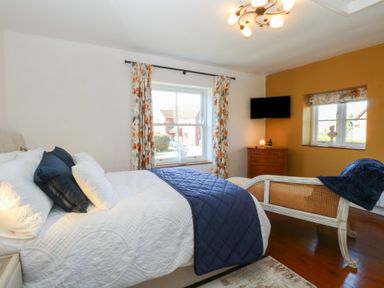 A bedroom with a bed white and blue bedding floral curtains a wooden dresser a wall-mounted TV and a bench at Cobble Cottage in Great Ryburgh