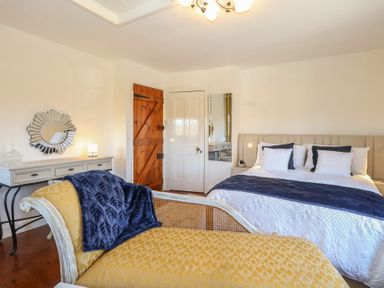 A bedroom with a beige bed and white bedding a yellow chaise lounge a wooden door and a white dresser at Cobble Cottage Great Ryburgh