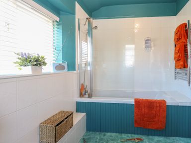 A bathroom with a bathtub and shower orange towels a plant and a radio on the windowsill at Cobble Cottage Great Ryburgh