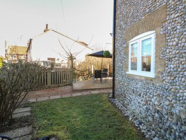A garden with grass and bushes next to a stone house with a wooden patio and outdoor furniture at Cobble Cottage in Great Ryburgh