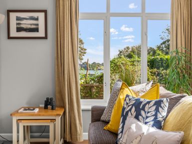 A room with a brown sofa with yellow and patterned pillows beside a window with beige curtains and a wooden side table with binoculars at Seascape in Kingsand