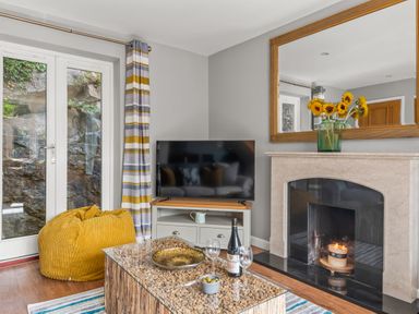 A living room with a television on a stand a fireplace with a candle and sunflowers in a vase on the mantle at Seascape in Kingsand