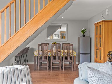 A dining area under wooden stairs with dark wooden chairs a table with a yellow patterned tablecloth a fridge a wooden cabinet and part of a sofa with pillows at Seascape in Kingsand
