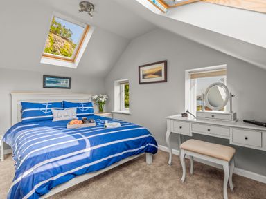 A bedroom with a blue and white striped bedspread a white dressing table with a mirror and a beige stool at Seascape in Kingsand